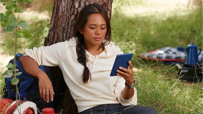 a person sits outside, under a tree while reading on a kindle device