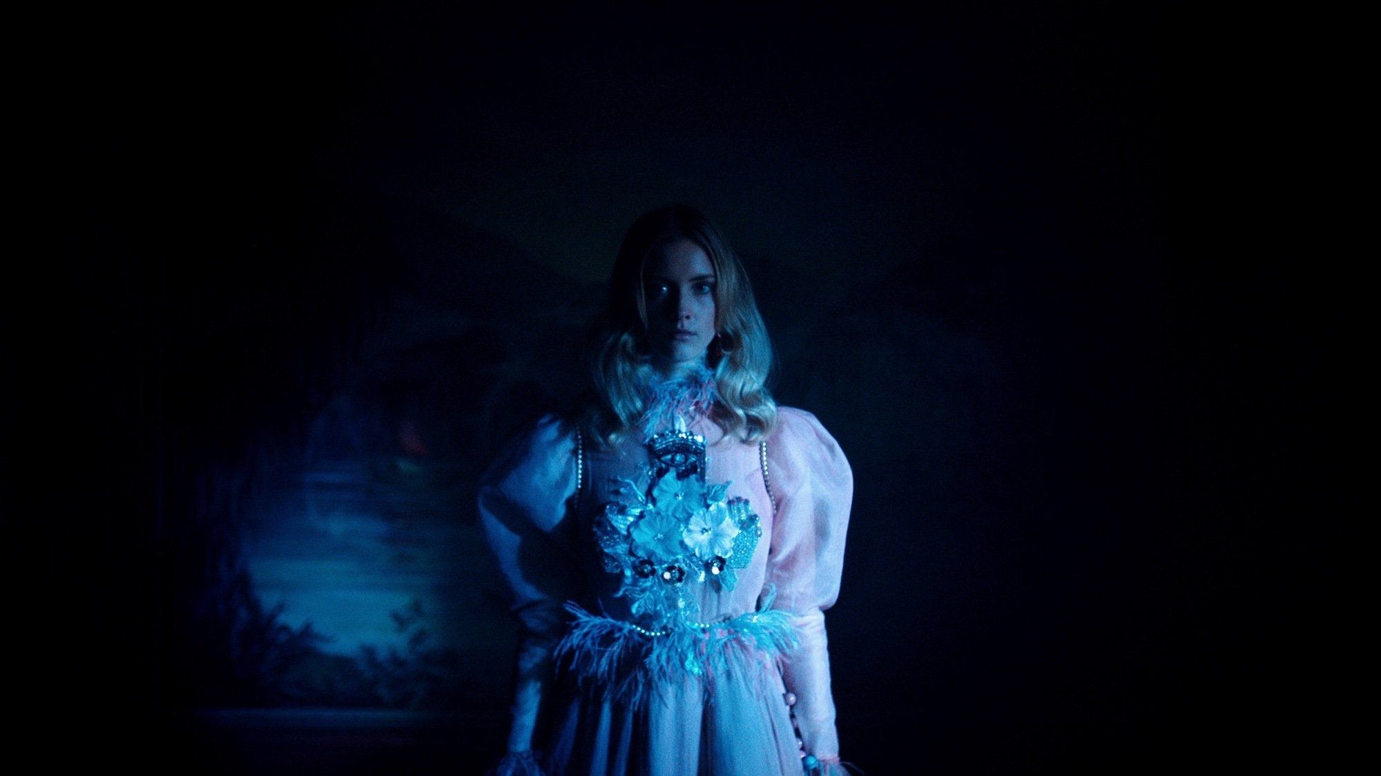 A blonde woman in a pink dress stands in a dark room. 