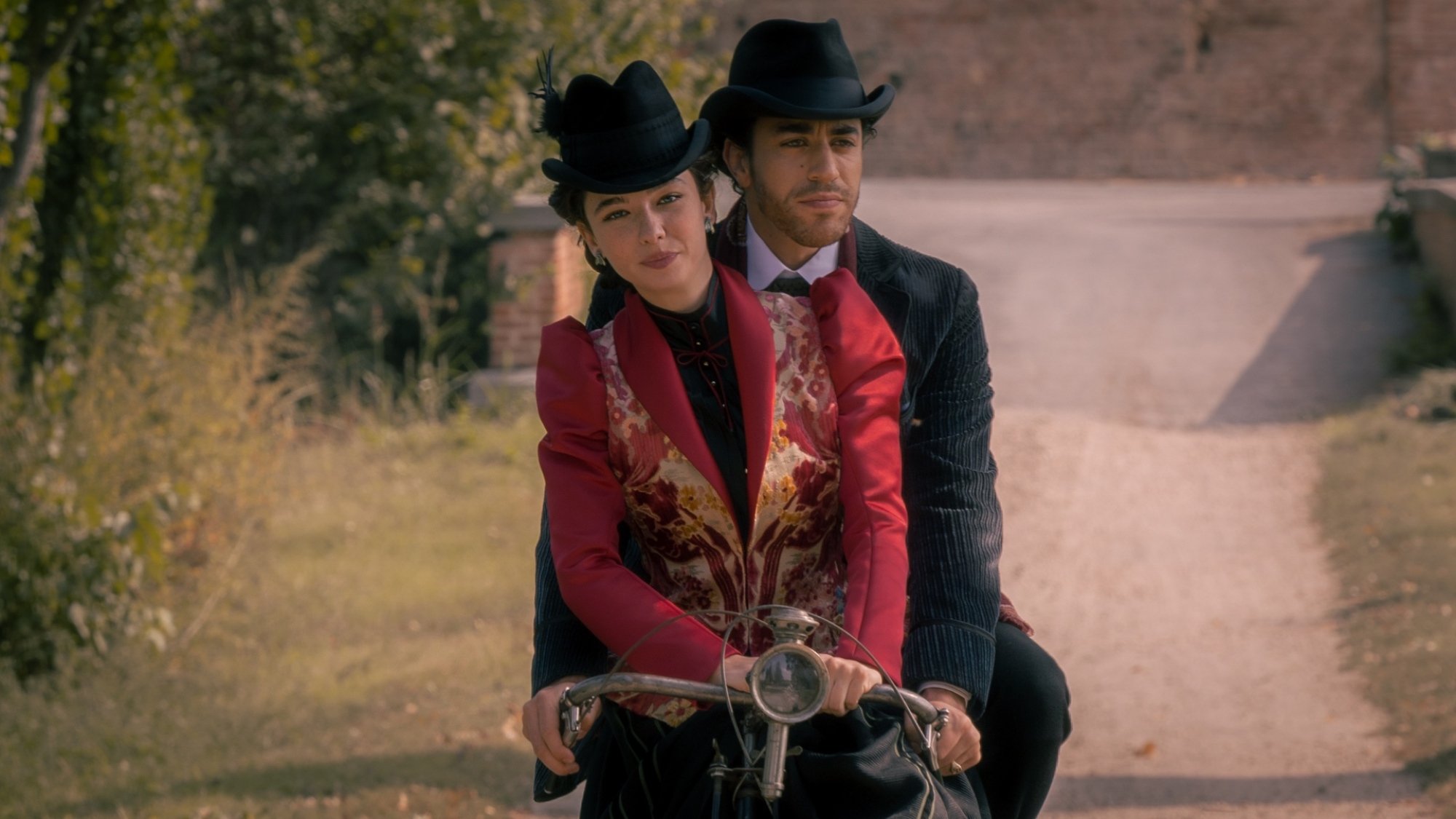 A woman in an old-fashioned red suit and black hat rides on the front of a bicycle driven by a man in a suit and hat.