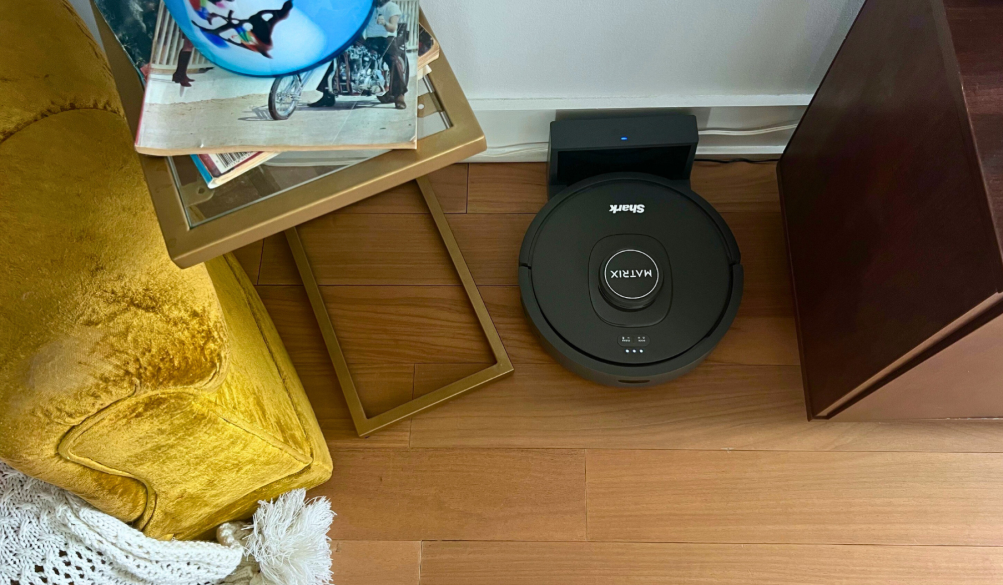 Shark RV2300 robot vacuum on dock against wall with yellow armchair and TV stand in peripherals