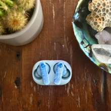 Skullcandy EcoBuds headphones in case laying on wooden coffee table with potted cactus and seashell decor in peripherals