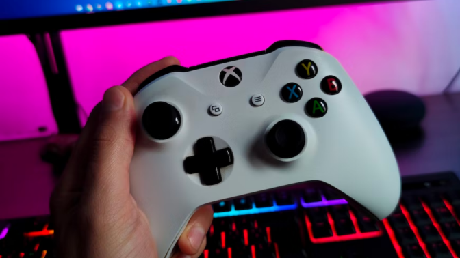 a close-up of a person holding an xbox controller in front of.an RGB keyboard and a gaming monitor