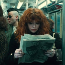 A woman with voluminous, curly red hair reading a newspaper on a public train; still from "Russian Doll" Season 2.