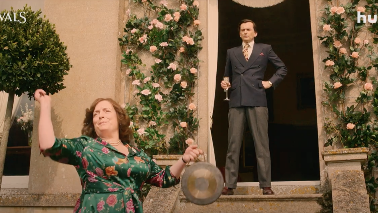 A woman bashes a gong while a man in a suit stands in a doorway with a flute of bubbly.