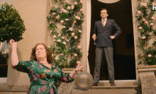 A woman bashes a gong while a man in a suit stands in a doorway with a flute of bubbly.