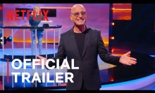 Still of a bald man with his hands outstretched on a game show stage.