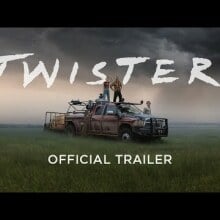 A man and woman standing on top of a truck in a field under stormy skies.