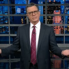 A man in a suit stands on a talk show stage, gesturing with his arms wide.