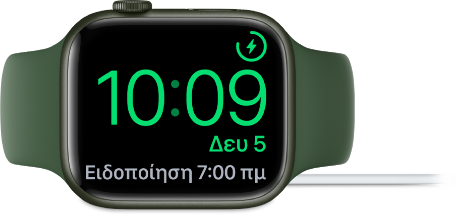 Ένα Apple Watch τοποθετημένο στο πλάι του και συνδεδεμένο στον φορτιστή του. Στην οθόνη εμφανίζεται το σύμβολο φόρτισης στην πάνω δεξιά γωνία, η τρέχουσα ώρα από κάτω η ώρα του επόμενου ξυπνητηριού.