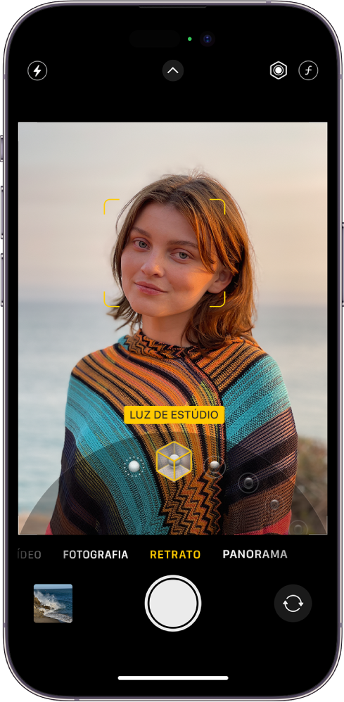 Ecrã da Câmara no modo Retrato; no visor, o objeto está nítido e o fundo desfocado. O seletor para escolher os efeitos de iluminação está aberto na parte inferior da moldura e a opção “Luz de estúdio” está selecionada. Na parte superior do ecrã, à esquerda, encontra‑se o botão do flash, na parte superior encontra-se o botão de controlos da câmara e, em cima à direita, encontram‑se os botões para ajustar a intensidade dos efeitos de iluminação e o controlo de profundidade. Na parte inferior do ecrã encontram-se, da esquerda para a direita, o botão de visualização de fotografias e vídeos, o botão para tirar fotografia e o botão "Seletor da câmara - traseira”.