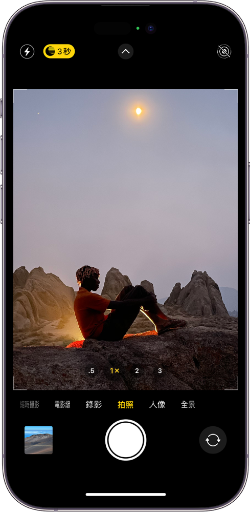 「拍照」模式的「相機」螢幕。觀景窗中顯示夜景。閃光燈和「夜間」模式按鈕顯示在螢幕的左上角，「夜間」模式已開啟。「相機控制項目」按鈕位於最上方中央，「原況照片」按鈕位於右上角。螢幕底部由左至右為「照片和影片檢視器」按鈕、「拍照」按鈕和「相機選擇器後置」按鈕。