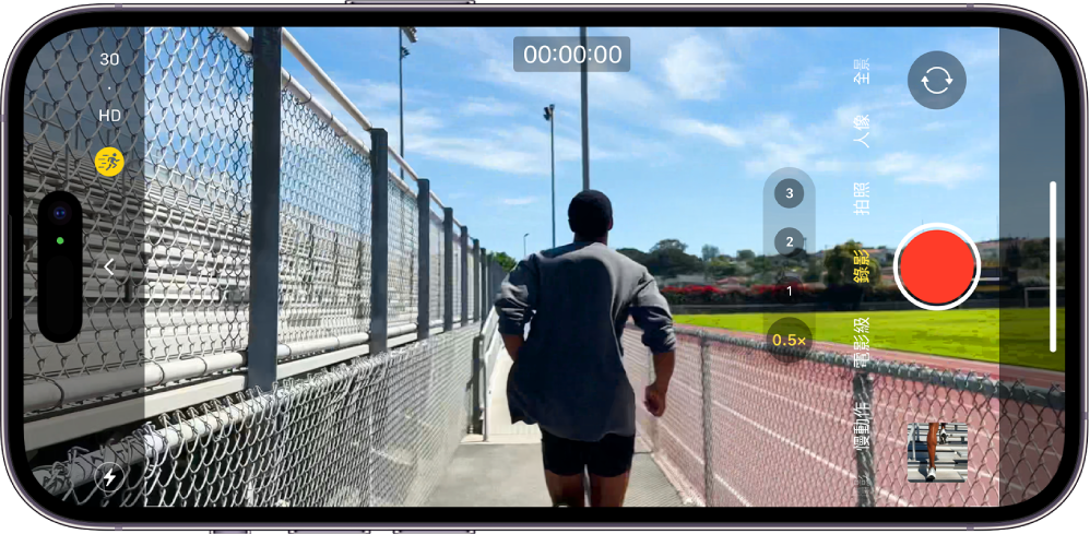 將 iPhone 旋轉為橫向。「相機」以「錄影」模式打開。在相機觀景窗中，一個人正在慢跑。「閃光燈」按鈕位於螢幕左下角。「相機控制項目」按鈕位於螢幕左側中央。左上角是快速切換，可切換影片解析度和影格率。左上角的「動作」按鈕已開啟。右側由上至下為「相機選擇器後置」按鈕、「錄影」按鈕，以及「照片和影片檢視器」按鈕。觀景窗中顯示一個奔跑中的人。