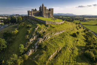 A castle stands on top of a rocky, grassy cliff reaching towards the sky