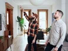 Two Men Stand in Living Room, One Pointing at Ceiling in New York Home