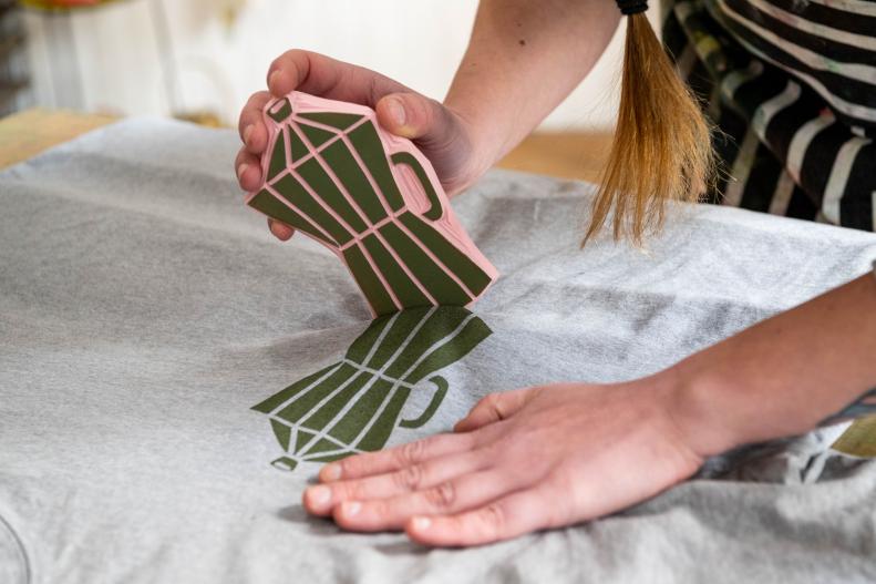 Woman Uses Hand to Stamp Green Print of Coffee Pot Onto Shirt