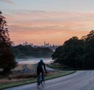 an unrecognisable cyclist on a winding road at dawn in a london park city of london in the background