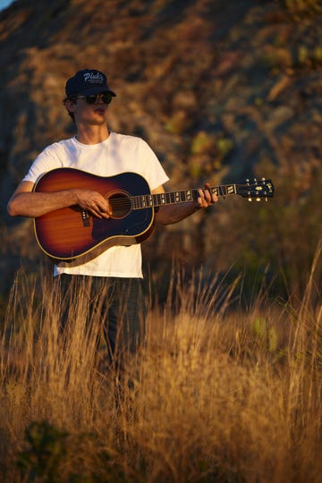 calder allen holding guitar, standing in field of tall grass wearing baseball cap, sunglasses, white tee shirt, jeans