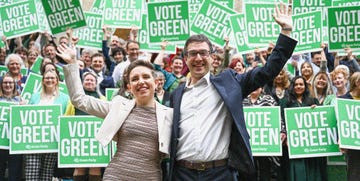 carla denyer and adrian ramsay stand in front of supporters holding signs saying vote green