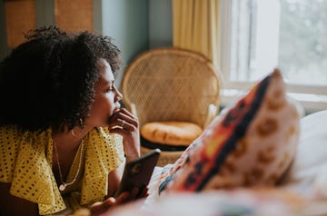 a woman looks as though she is reflecting on life as she gazes out of a bedroom window she lies on her stomach on a bed, and touches her chin with her hand she is holding a smart phone but isnt looking at it