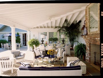 a large outdoor room with a large patio and a large patio with white chairs and brick fireplace and plant in the background