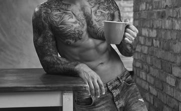 handsome bearded tattoed male with a stylish haircut and muscular body, drinks coffee, leaning on a table