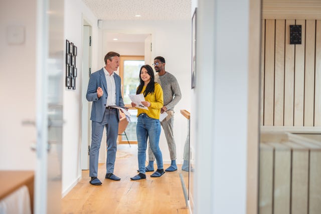 an adult caucasian male real estate agent giving an apartment tour to a young diverse couple the real estate agent is wearing a fancy suit while the couple is wearing casual clothes together they are walking through the hall of the apartment the rooms have modern decor with wooden details