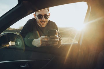 handsome young man using smart phone while leaning on car window during sunset