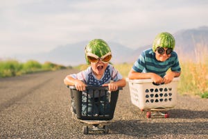 a young boy races his brother in a makeshift go cart while wearing watermelon helmets and goggles he is excited as he is winning the race