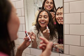 smiling female friends applying lipstick while reflecting in mirror at home