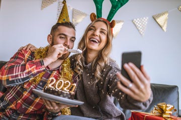 couple celebrating christmas at home, holding chocolate cake with lit candles shaped as numbers 2021 and having a video call with friends