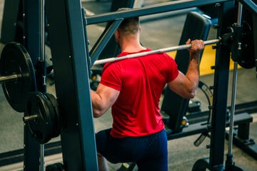 rear view of muscular male athlete doing weighted lunges in front of a mirror at the gym