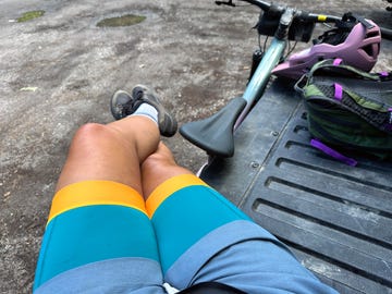 a person sits on a bench in bike shorts next to bike, helmet, backpack