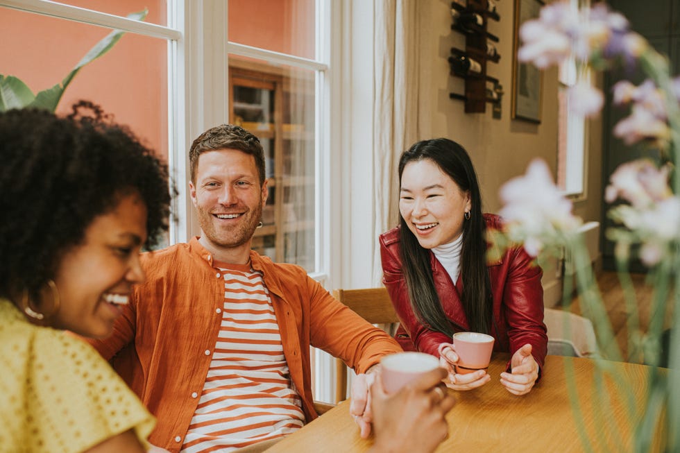 three young people socialise at a table while enjoying hot drinks they are animated as they giggle together one girl is in the foreground whilst focus remains on a smiling male and female