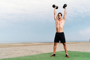 young muscular man training shoulder lifts on the beach shirtless lifting dumbbells