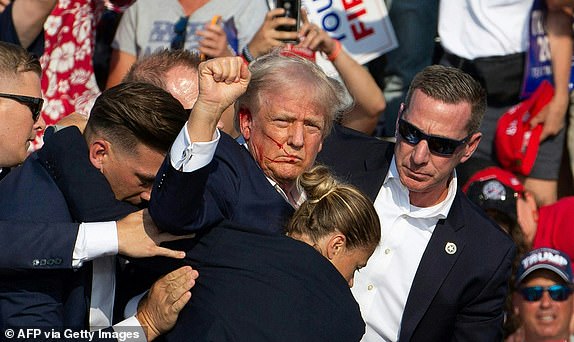 TOPSHOT - Republican candidate Donald Trump is seen with blood on his face surrounded by secret service agents as he is taken off the stage at a campaign event at Butler Farm Show Inc. in Butler, Pennsylvania, July 13, 2024. Donald Trump was hit in the ear in an apparent assassination attempt by a gunman at a campaign rally on Saturday, in a chaotic and shocking incident that will fuel fears of instability ahead of the 2024 US presidential election. The 78-year-old former president was rushed off stage with blood smeared across his face after the shooting in Butler, Pennsylvania, while the gunman and a bystander were killed and two spectators critically injured. (Photo by Rebecca DROKE / AFP) (Photo by REBECCA DROKE/AFP via Getty Images)