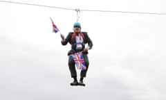 Boris Johnson hanging on a zip wire holding union jack flags over a park, with a tower block in the background