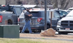 A white man wearing a hat, sunglasses, jeans and bulletproof vest walks through a parking lot carrying a machine gun pointed at the ground.