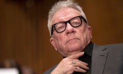 Older white man with short gray hair with black-framed glasses wearing a suit, with what looks like the hold 'HOLD' tattooed across the knuckles of his right hand.