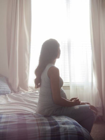 A woman sitting on a bed looks outside of a glowing window.