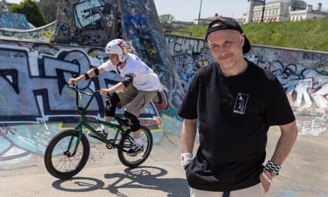 A young man stands posing for a picture in a skate park while a child rides past behind him on a BMX bike