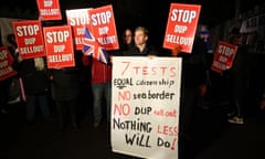 A group of protesters pictured at night, holding red signs bearing the words 'Stop DUP sellout'. One protester also holds a Union Jack. Another man in the foreground holds a large placard that reads: 'Seven tests, equal  citizenship, no sea border, no DUP  sellout, nothing less will do!'