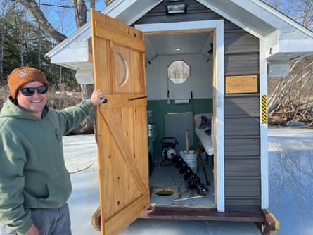 smiling man holds open door of small house to show small room with cot and equipment