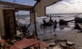 A view from inside the remains of a building, with the roof and much of the walls missing, of uprooted trees and a boy running out of the waves on the shoreline