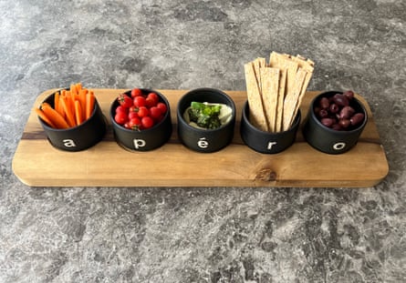 Five small black dish, each stamped with a letter so when they’re lined up they spell “apéro”. They are filled with an assortment of snacks including carrot sticks, cherry tomatoes, cheese, crackers and olives.