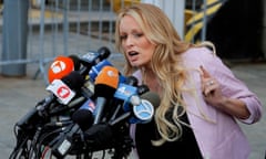 White woman, pink blazer, holds up finger and speaks into clutch of microphones.