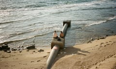 Two young men, topless and wearing shorts, sitting on a pipeline on a beach that leads into the sea