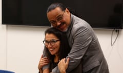 In a florescent-lit room with a massive flat-screen television on one wall, a Native woman with long brown hair and black cat's-eye glasses smiles broadly, sitting in a chair and holding the arms of a man who embraces her from behind. He is also Native, with wire-rimmed glasses and long, combed-back black hair, with a salt-and-pepper goatee and a gray blazer.