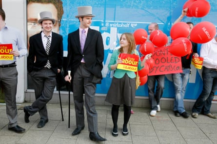 Two young men in suits and top hats with canes stand beside campaigners with Labour party paraphernalia, balloons and placards.