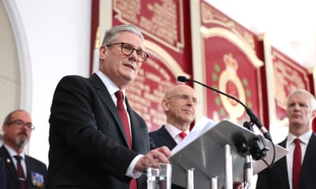 Keir Starmer speaking at the Fusilier museum in Bury: he is at a lectern behind a microphone, while three other men look on; they are all dressed in dark suits with red ties and stand in front of a large wall-mounted plaque in dark red and gold with military insignia