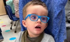 Toddler wearing blue glasses and a green scarf can be seen resting his head against a fabric headrest, mouth ajar and looking to the right